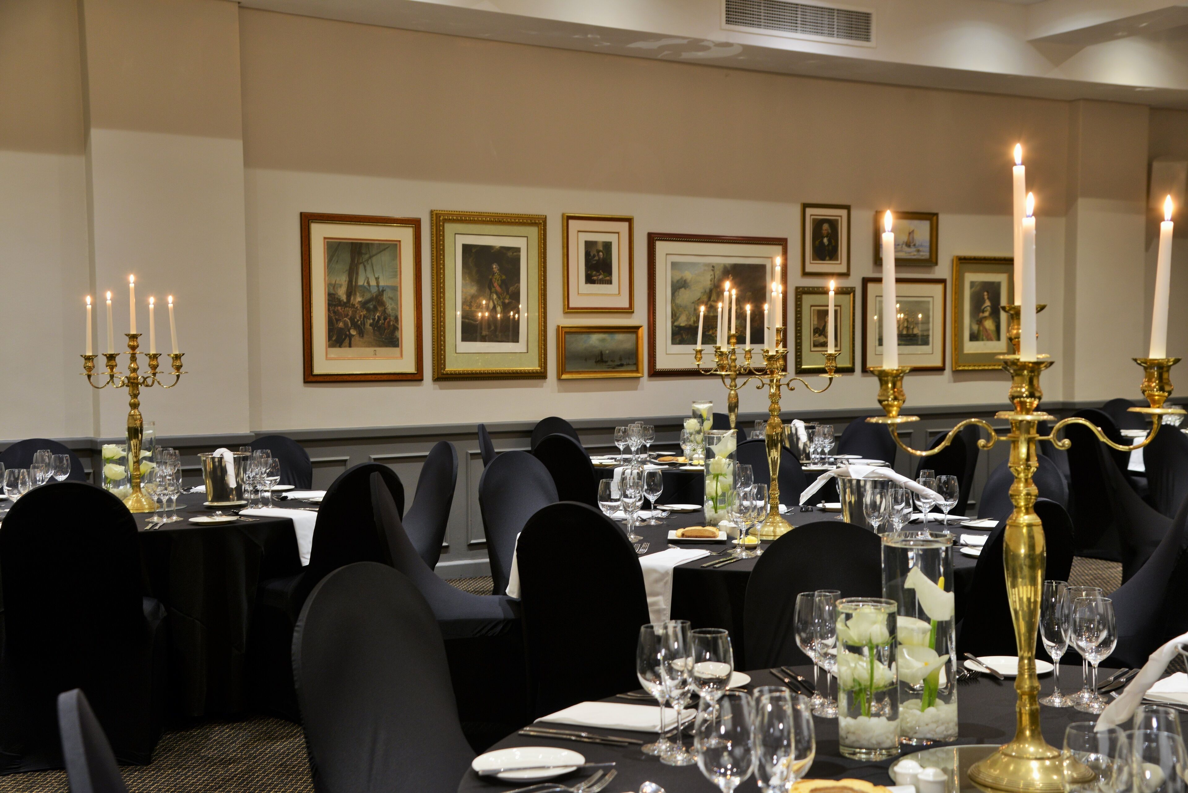 A formal dining room with tables set for a meal, framed pictures on the wall, and candelabras with lit candles.