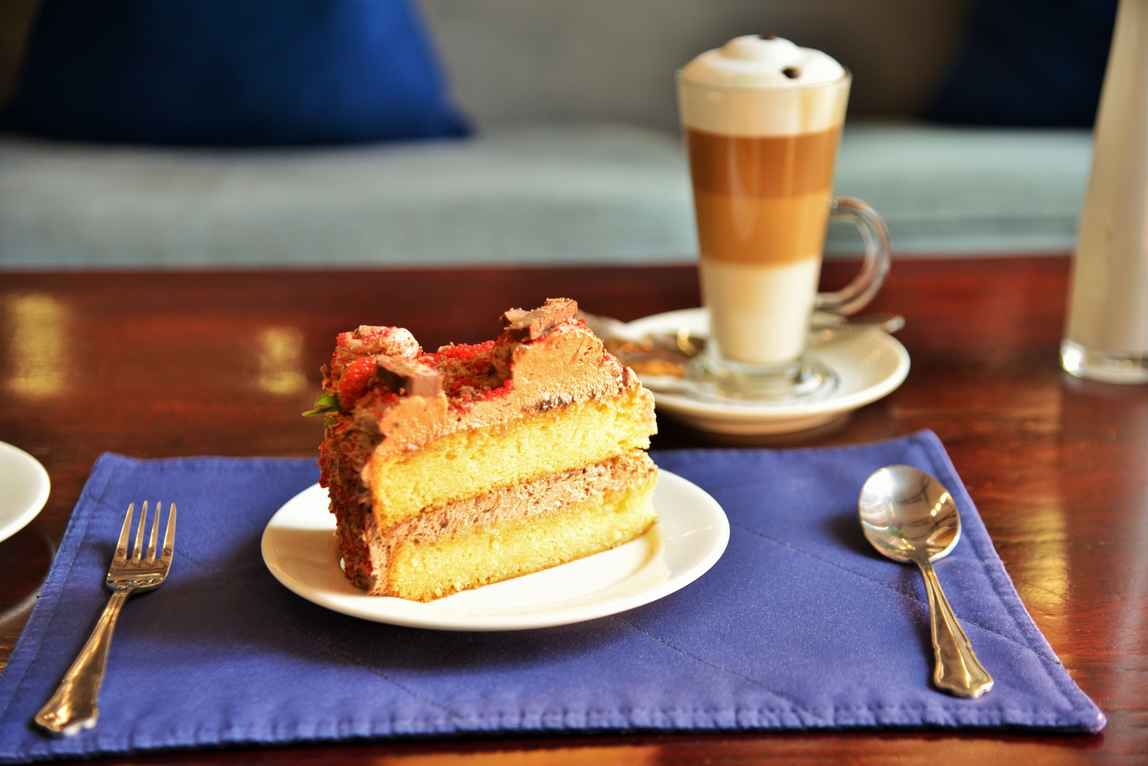 Une part de gâteau à plusieurs couches, présentée sur une assiette blanche, accompagnée d’une tasse de café et d’une cuillère posée sur une serviette bleue.