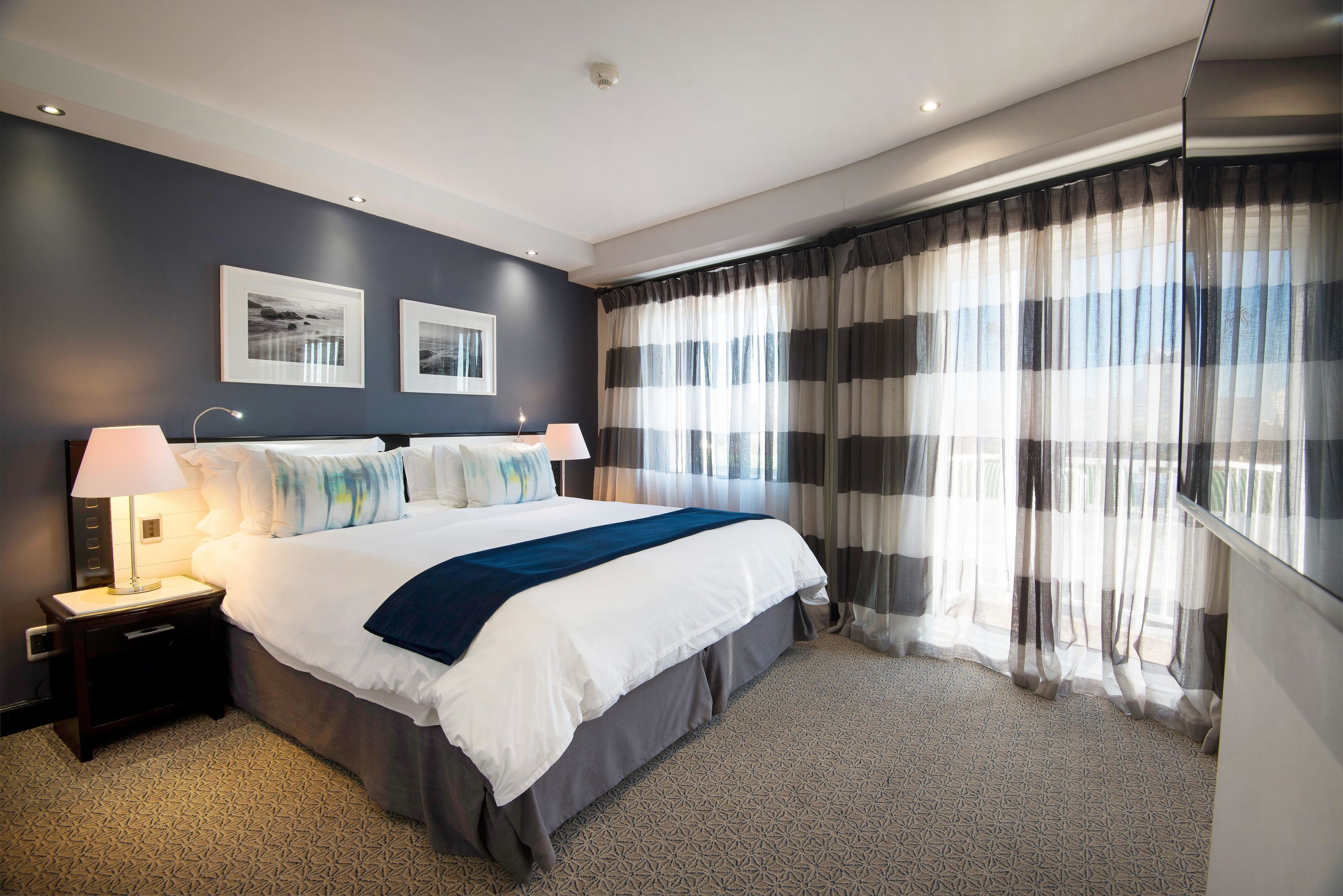 A modern hotel room with a large bed, two bedside tables with lamps, a patterned carpet, and a view through sheer curtains.