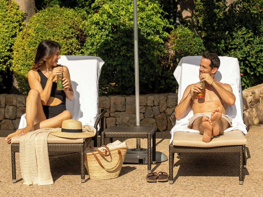 Two people relaxing on sun loungers with drinks, surrounded by greenery and a stone wall.