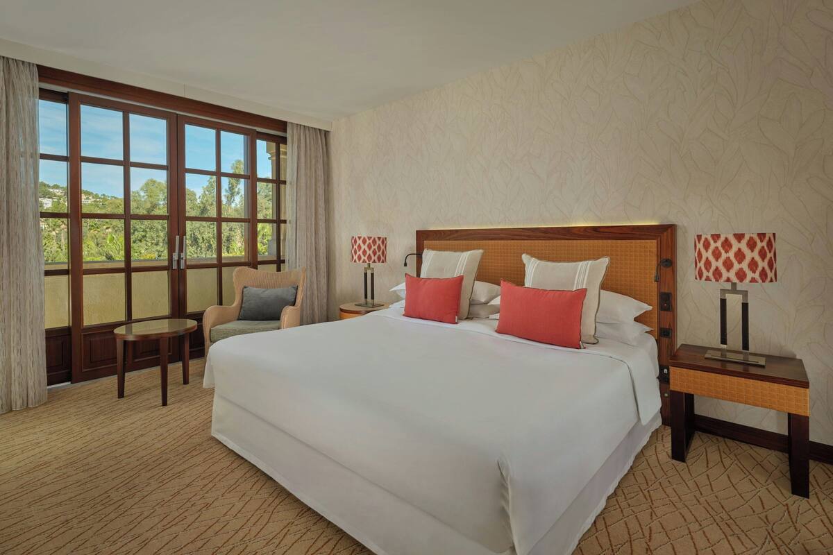 A hotel room with a large bed, two bedside tables with lamps, a small round table, a sofa, and a large window with a view of greenery.