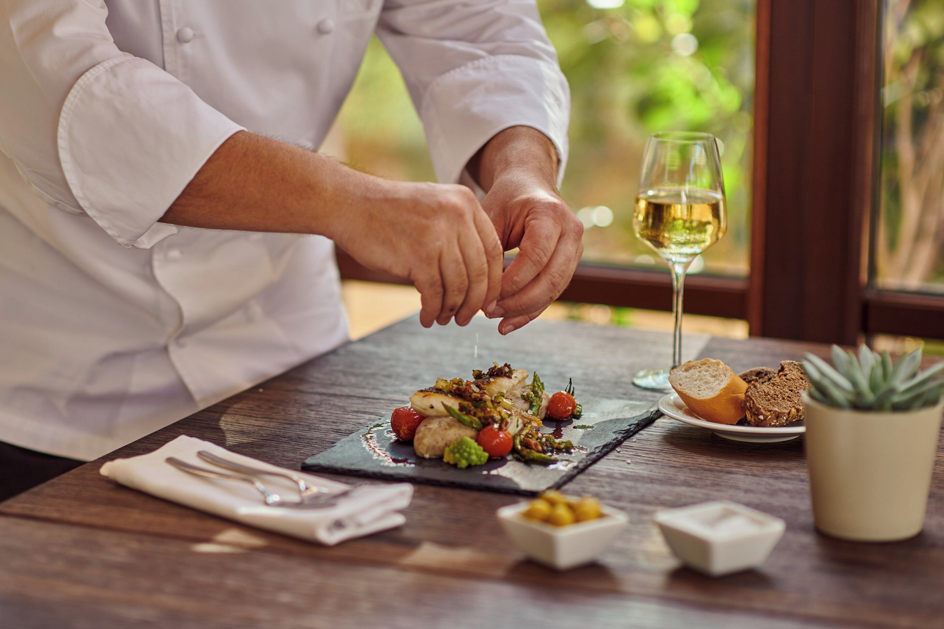 Un chef prepara un plato sobre una tabla de pizarra, con una copa de vino y pan sobre la mesa.