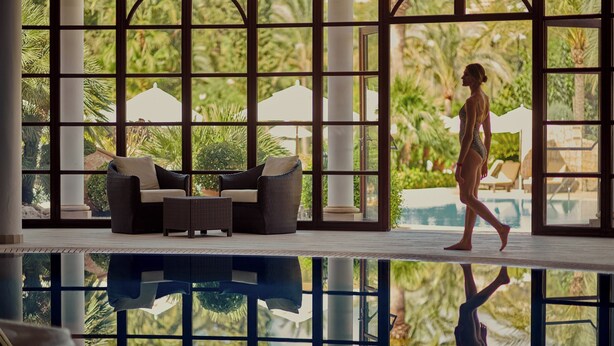Zona de piscina con piso de vidrio, mobiliario de mimbre y vistas a palmeras y sombrillas.