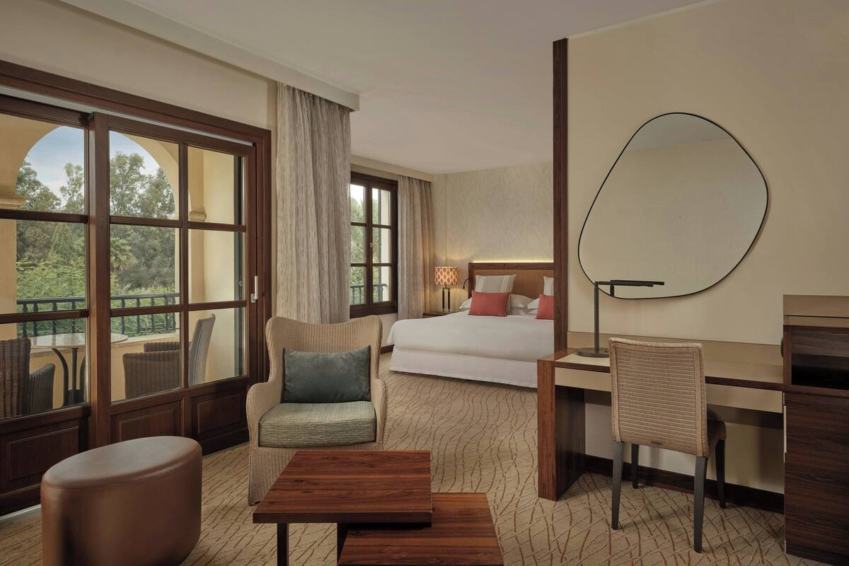 A hotel room with a large window, a bed, a desk, a chair, and a round mirror.