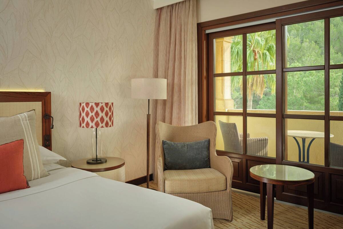 A hotel room with a bed, a chair, a table, and a large window with a view of palm trees.