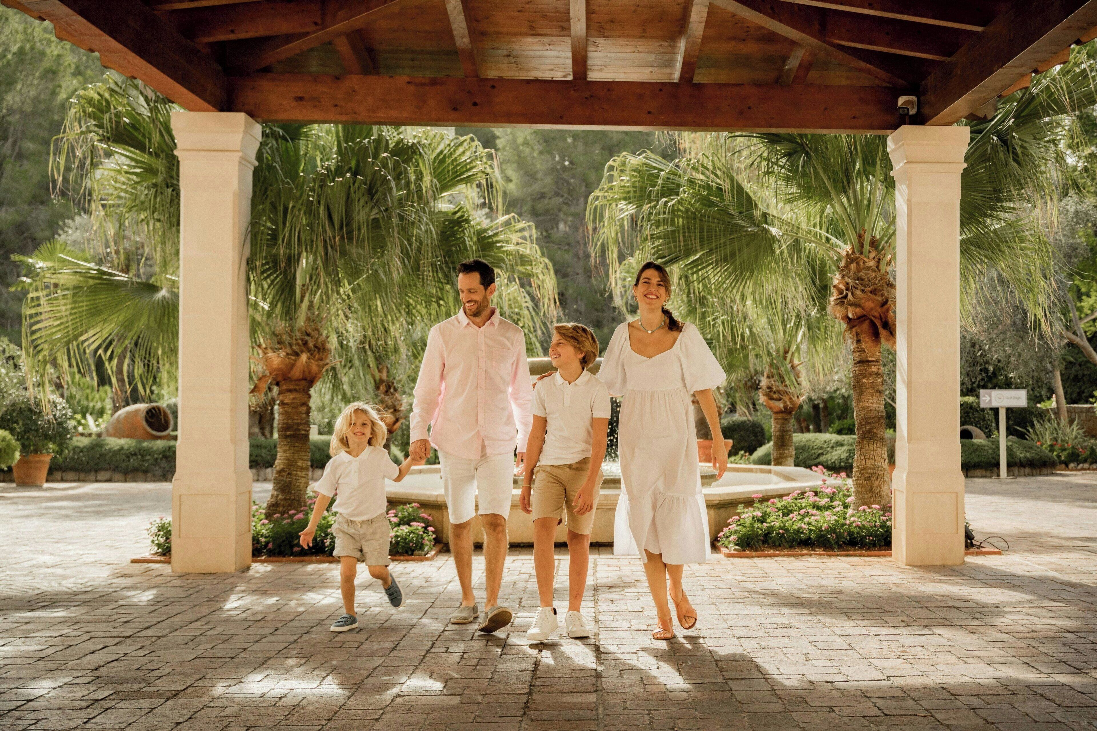 A family walking under a wooden pavilion with palm trees and a fountain in the background.