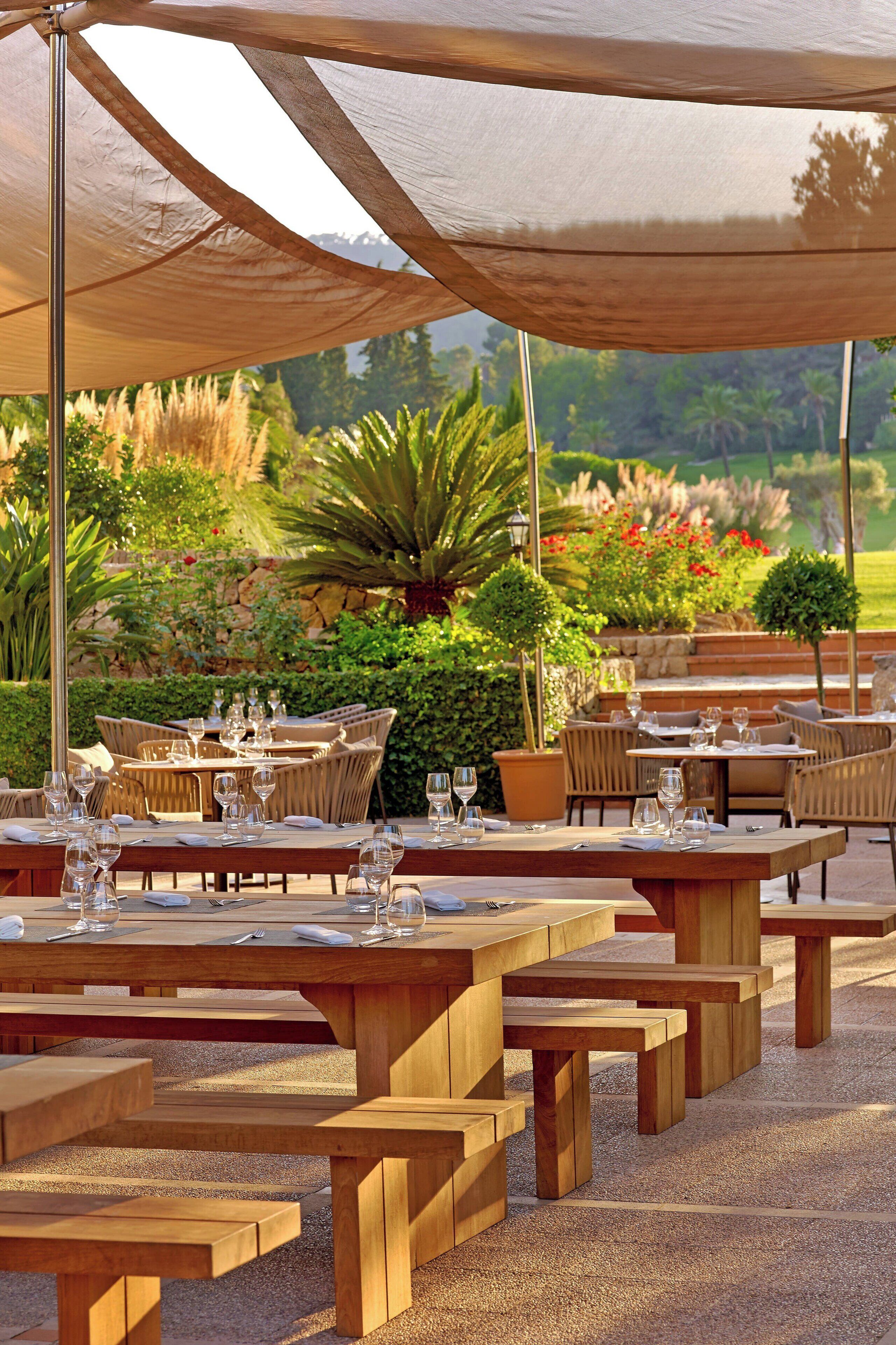 An outdoor dining area with wooden tables and chairs, shaded by large canopies, set against a backdrop of lush greenery and a stone wall.