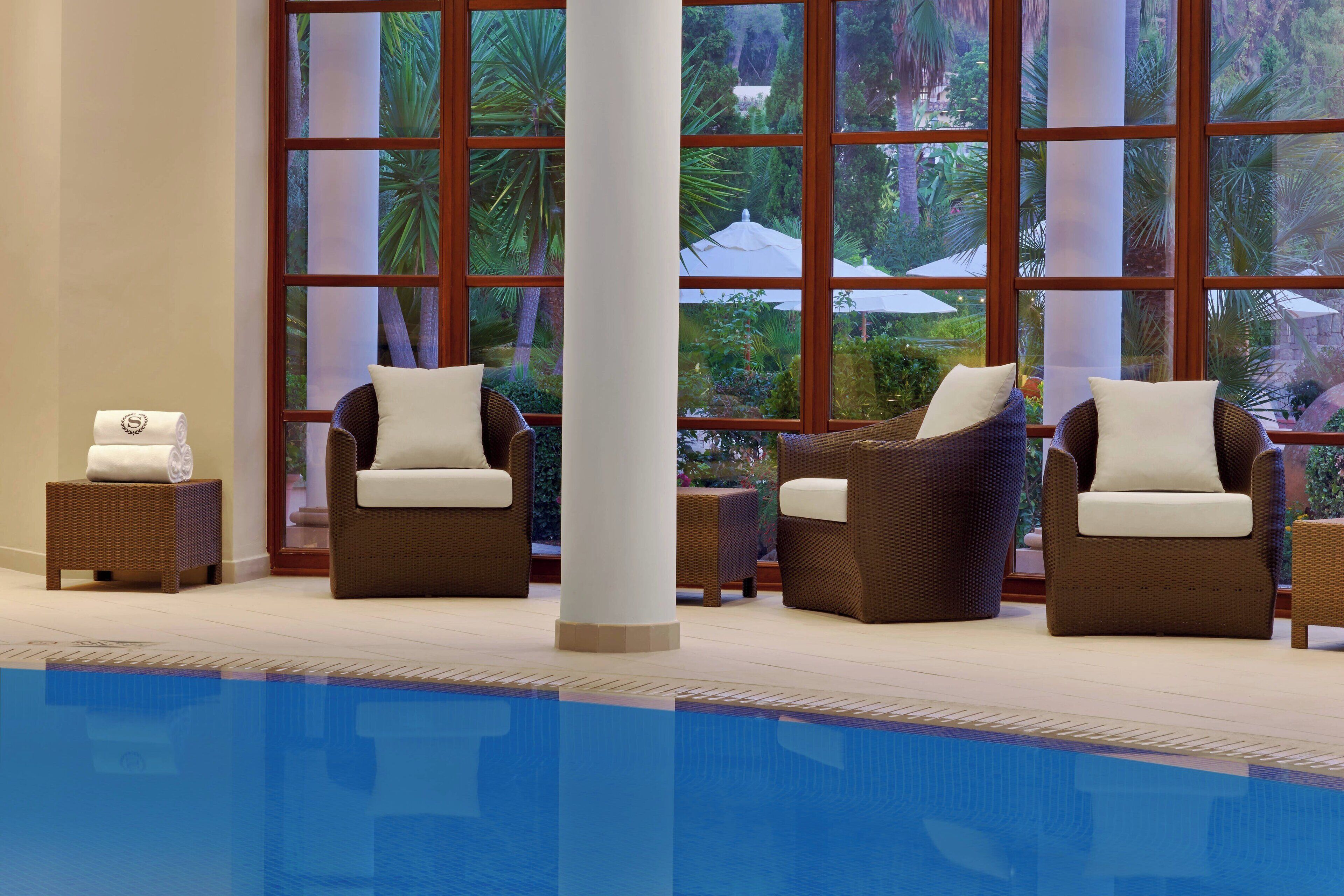 A poolside area with wicker chairs and a view of a tropical garden through large windows.