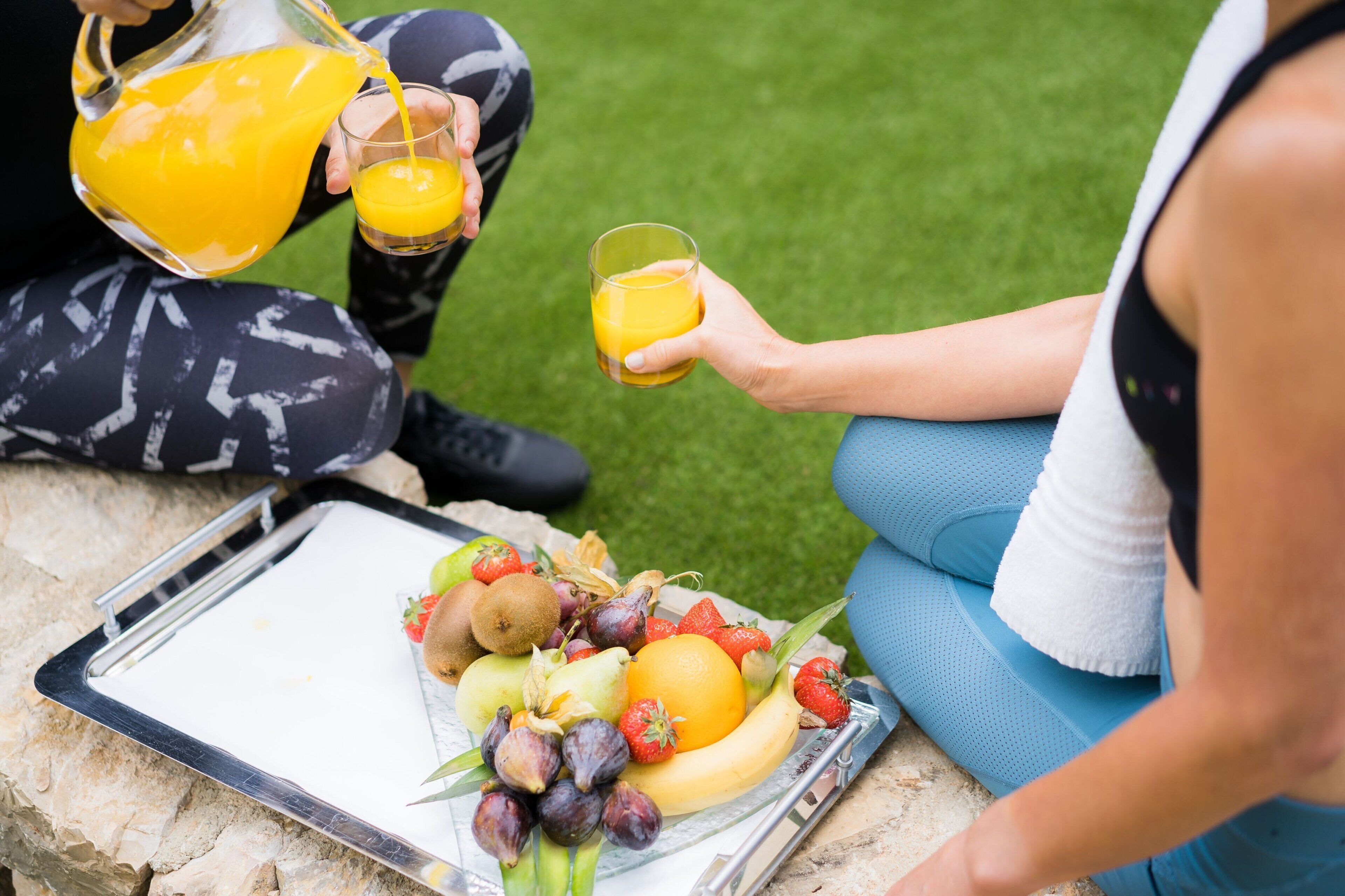 Una persona sirve jugo de naranja en un vaso, sentada en un banco de piedra con una bandeja de frutas y una toalla blanca cerca.