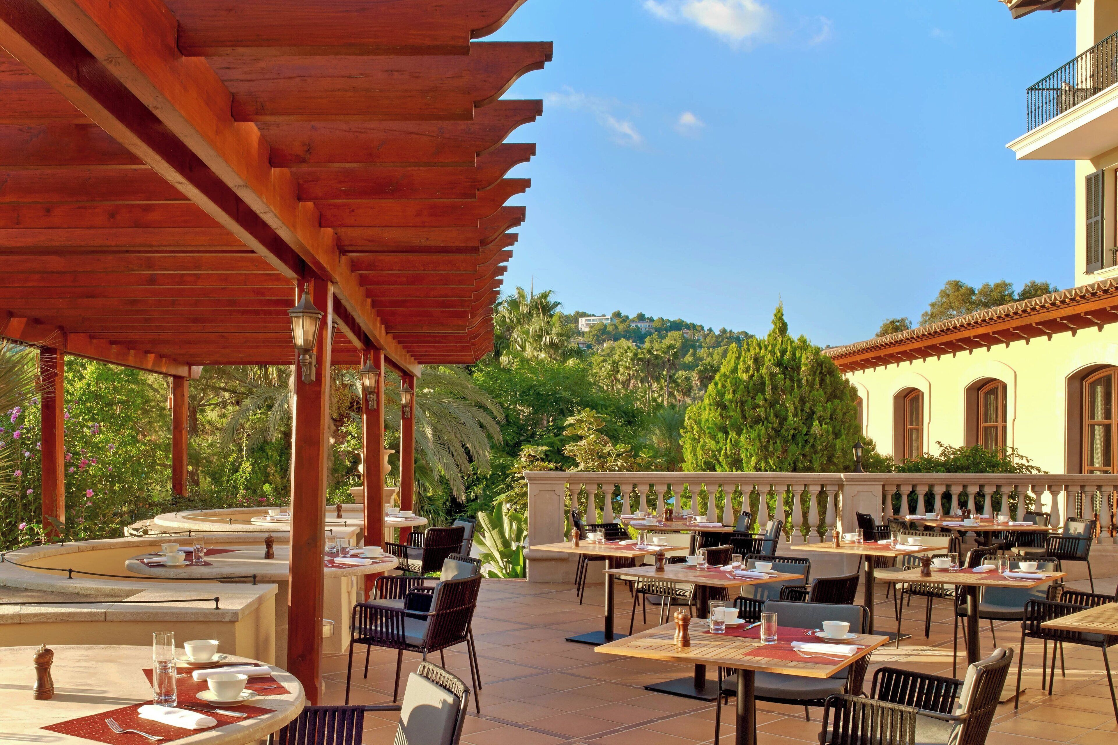An outdoor patio with tables and chairs, a wooden pergola, and a view of a hill with greenery and a building in the background.