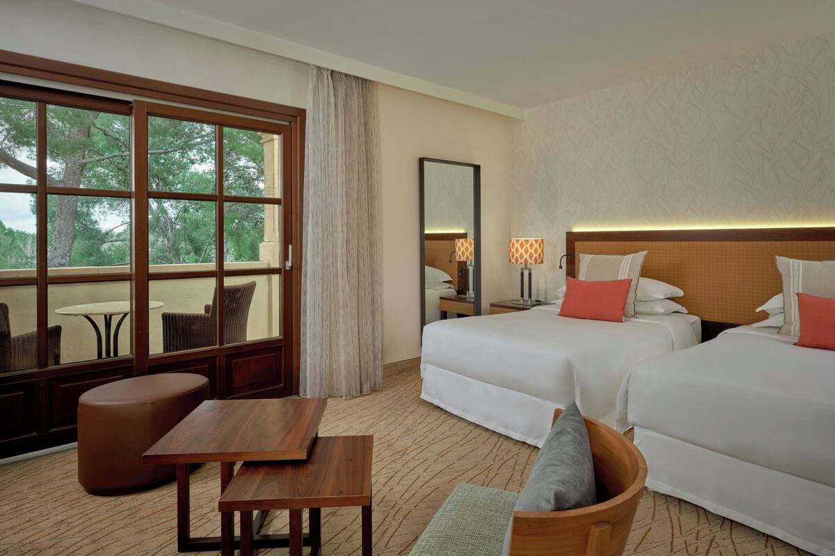 A hotel room with two beds, a wooden table, a small seating area, a large window with a view of trees, and a mirror on the wall.