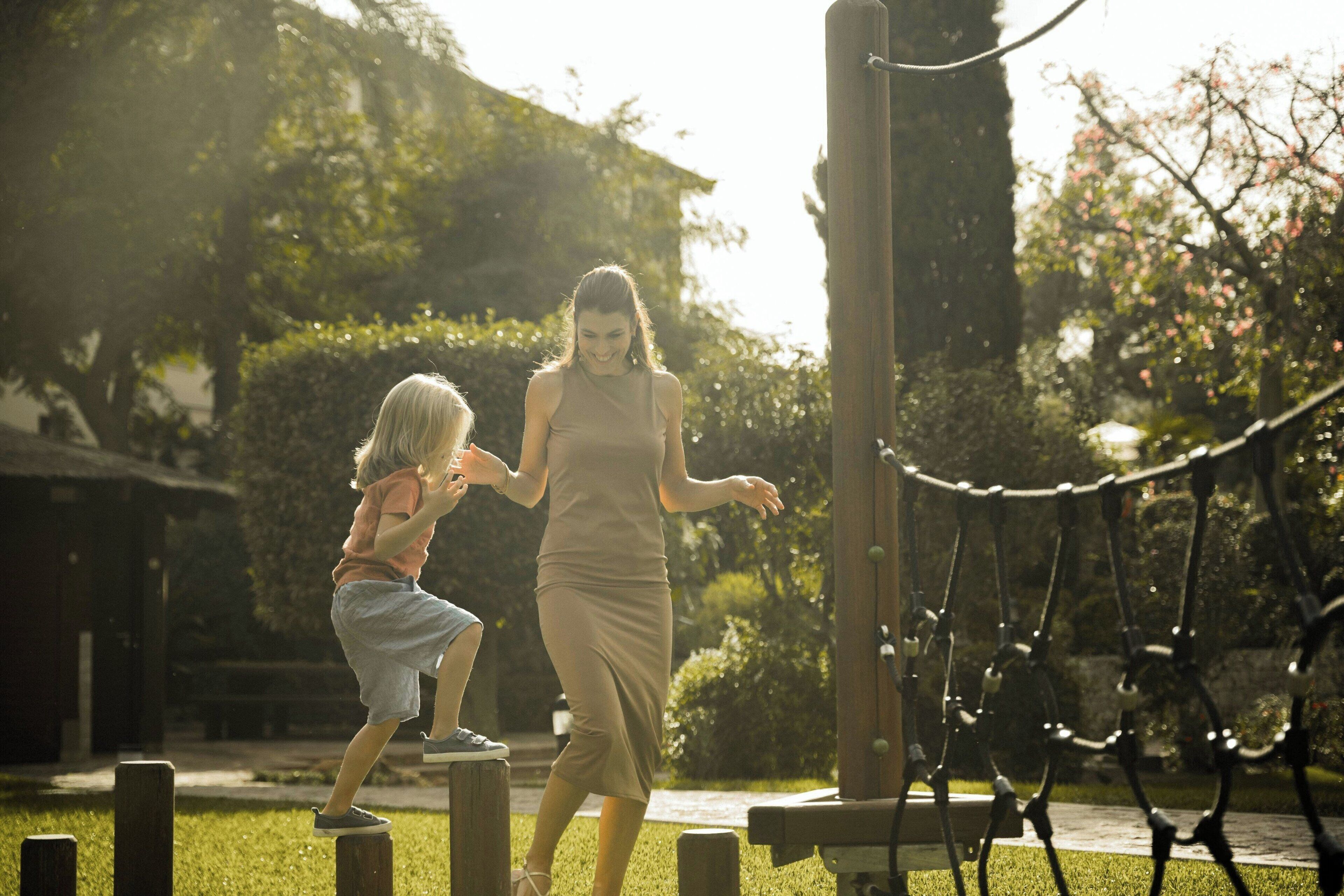 A woman and a child playing on wooden steps in a park with greenery and a wooden structure in the background.
