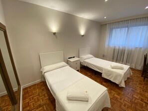 Ein Hotelzimmer mit zwei Einzelbetten, einem Holzboden mit Parkettoptik, einem kleinen Nachttisch mit Lampe und einem Fenster mit durchsichtigen Vorhängen.