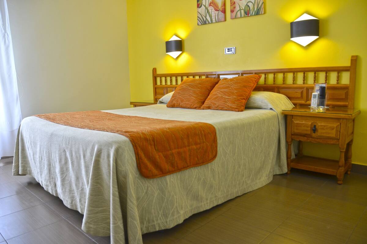 Uma cama com cabeceira de madeira, uma mesa de cabeceira com luminária e um candeeiro fixado na parede.
