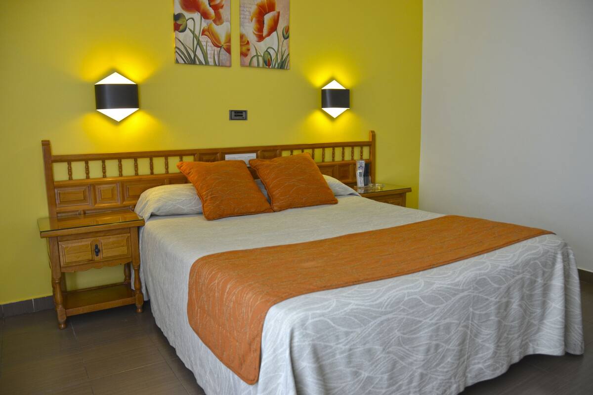 Uma cama bem arrumada com um cobertor laranja, duas luminárias fixadas na parede e uma mesa de cabeceira de madeira com gaveta.