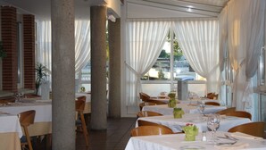 Un restaurante con mesas dispuestas para comer, ventanas amplias con cortinas y vistas al exterior.