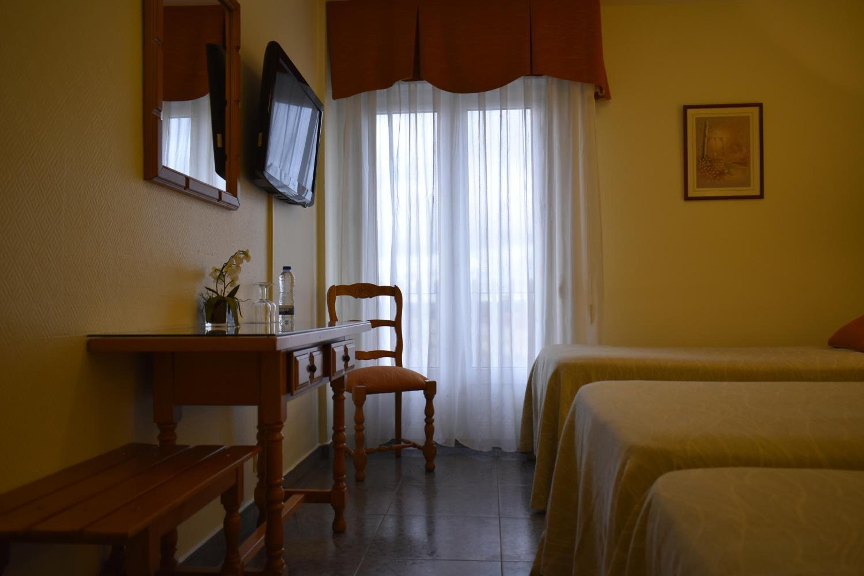 A hotel room with a wooden desk, a chair, a bed with a white bedspread, a television mounted on the wall, and a window with sheer curtains.