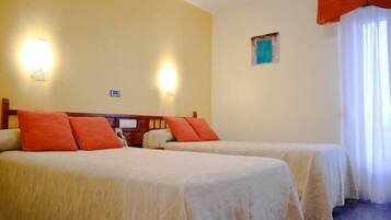 Ein Hotelzimmer mit zwei Betten, jeweils mit einer beigefarbenen Tagesdecke und orangefarbenen Kissen. An den Wänden sind Lampen angebracht und es gibt ein kleines Fenster mit Vorhängen.