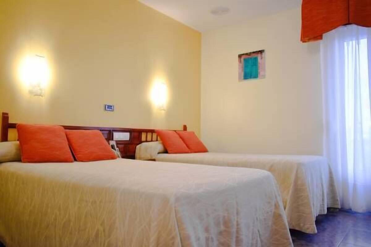 Quarto de hotel com duas camas, uma pequena janela e um candeeiro fixado na parede.