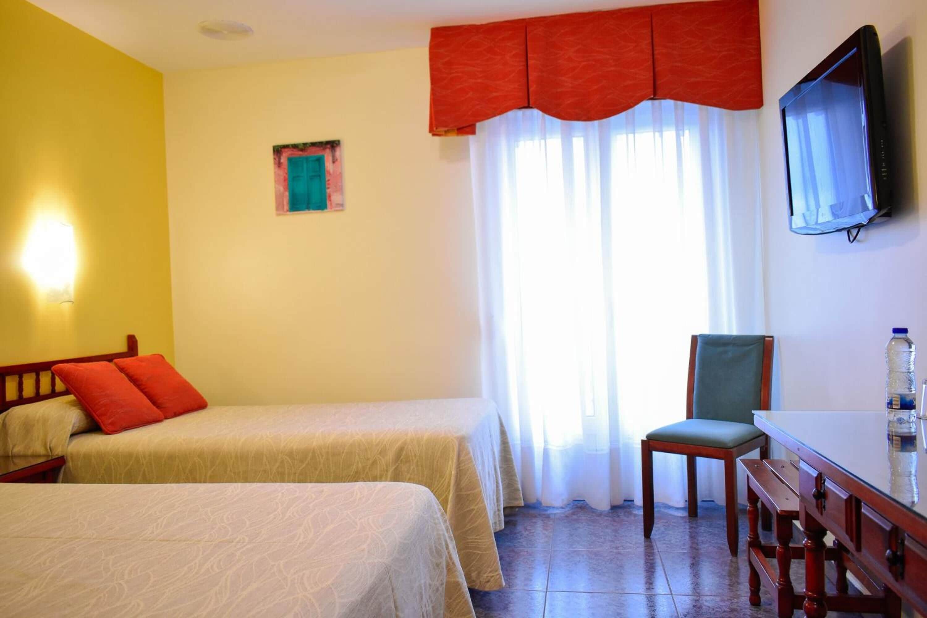 Habitación de hotel con dos camas, una silla, una mesa, un televisor y un ventanal con cortinas.