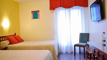 A hotel room with two beds, a chair, a table, a TV, and a window with curtains.