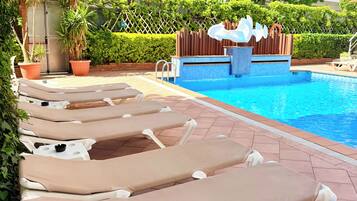 A hotel pool area with sun loungers, a pool, and a building with balconies.