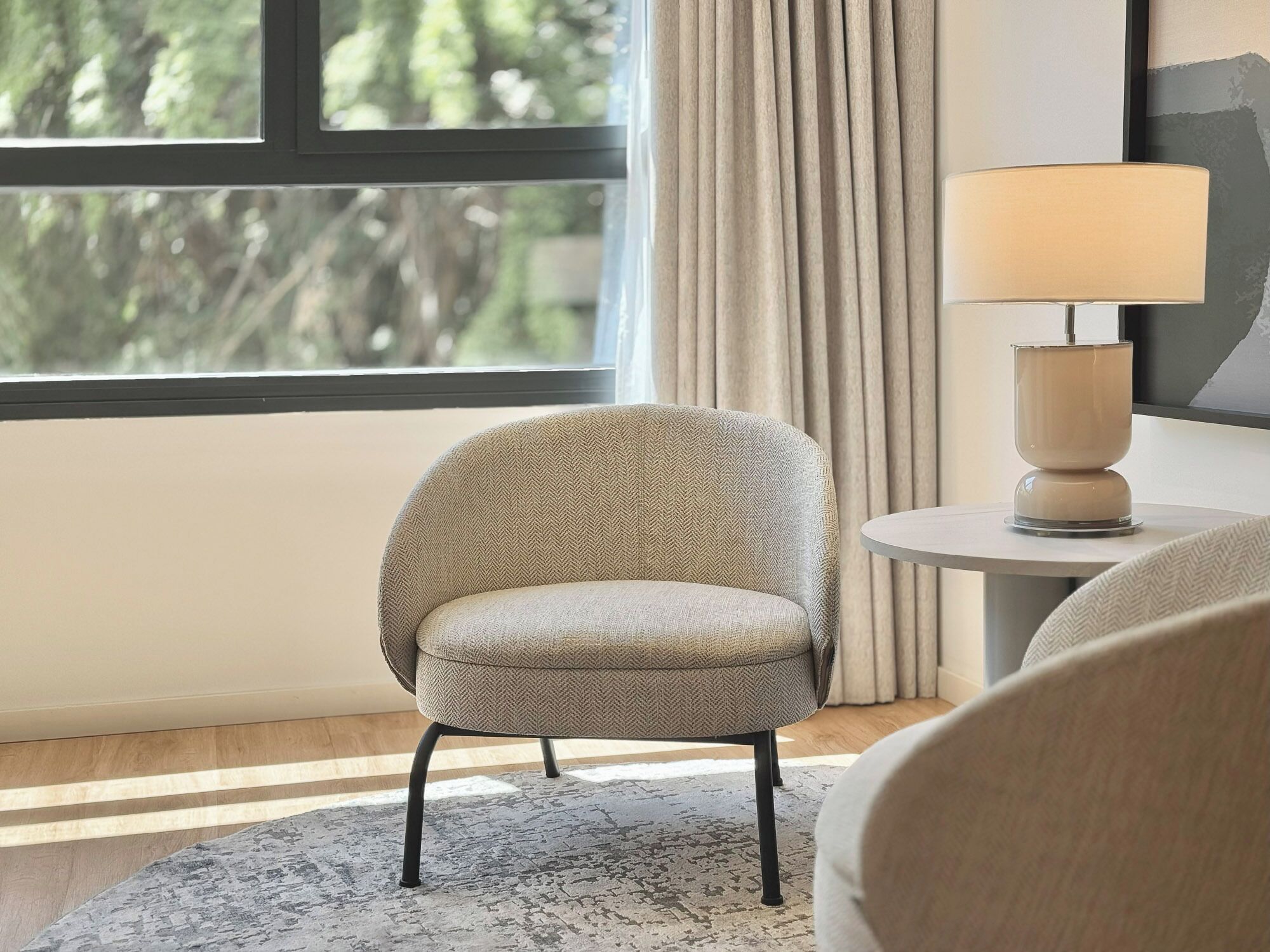 Una habitación moderna con un sillón de tapicería tipo espiga, una mesita redonda con una lámpara y vistas a árboles a través de una ventana con cortinas.