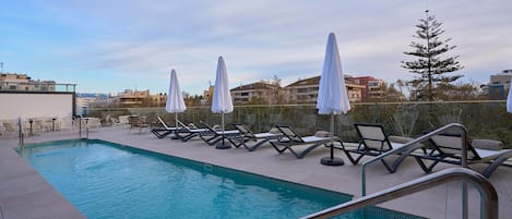A pool area with lounge chairs and umbrellas, surrounded by a fence and buildings in the background.