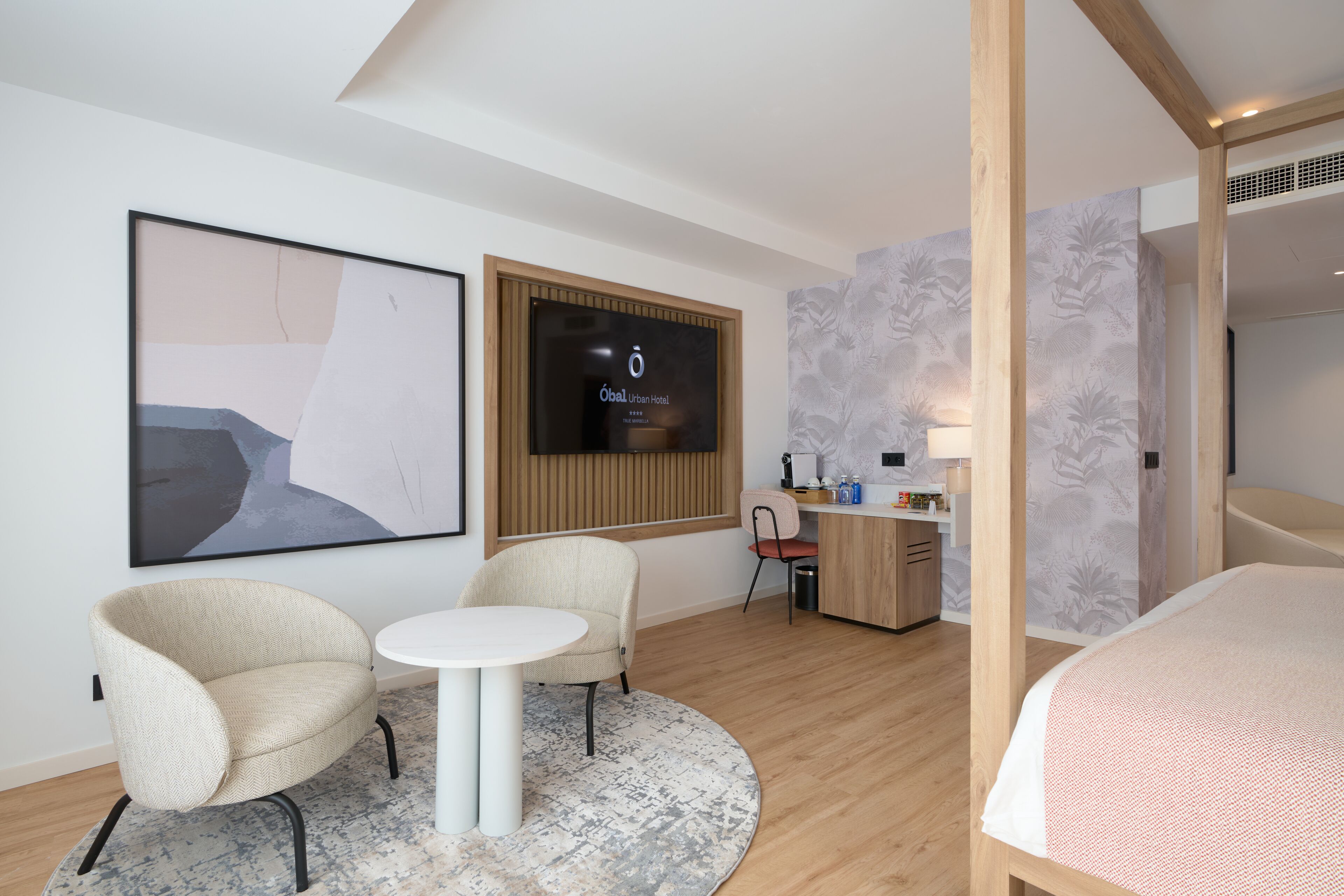 A modern hotel room with a bed, two armchairs, a small round table, a desk with a computer, and a TV displaying the hotel name.