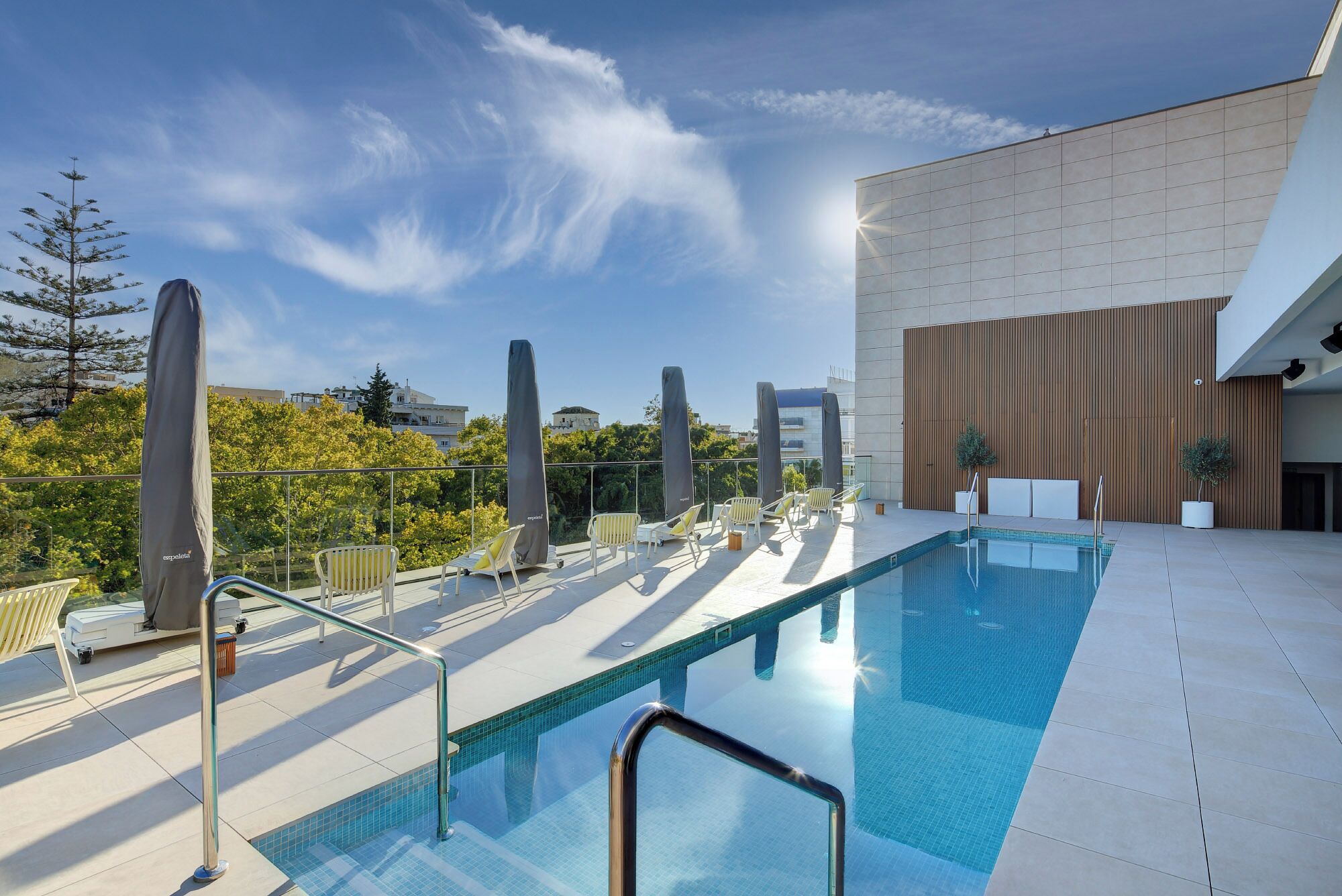 Una piscina en la azotea con sillones y sombrillas, rodeada por un edificio moderno con fachada de madera.