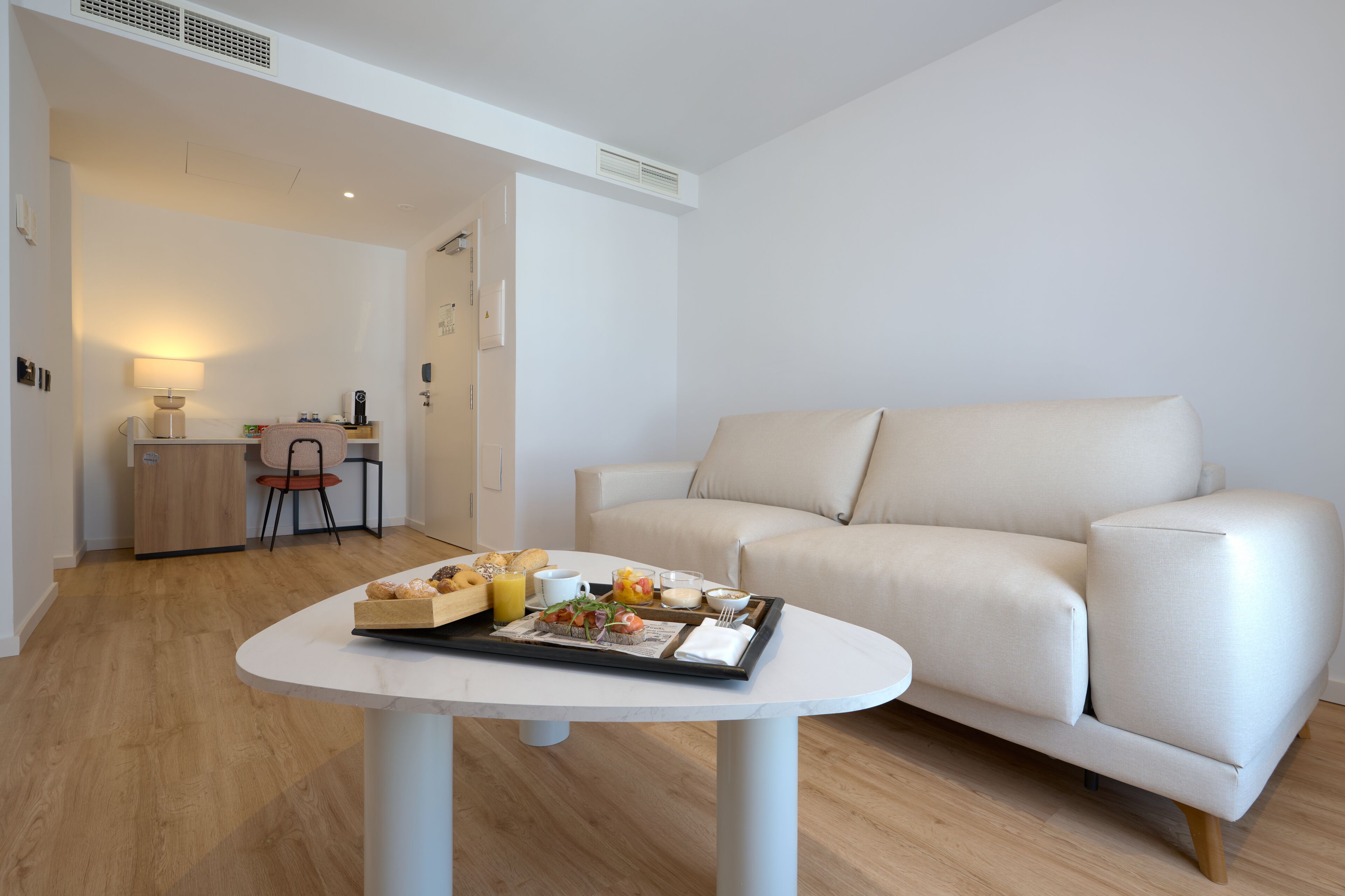 Een moderne woonkamer met een witte bank, een eettafel met ontbijtartikelen, en een klein keukenhoekje op de achtergrond.