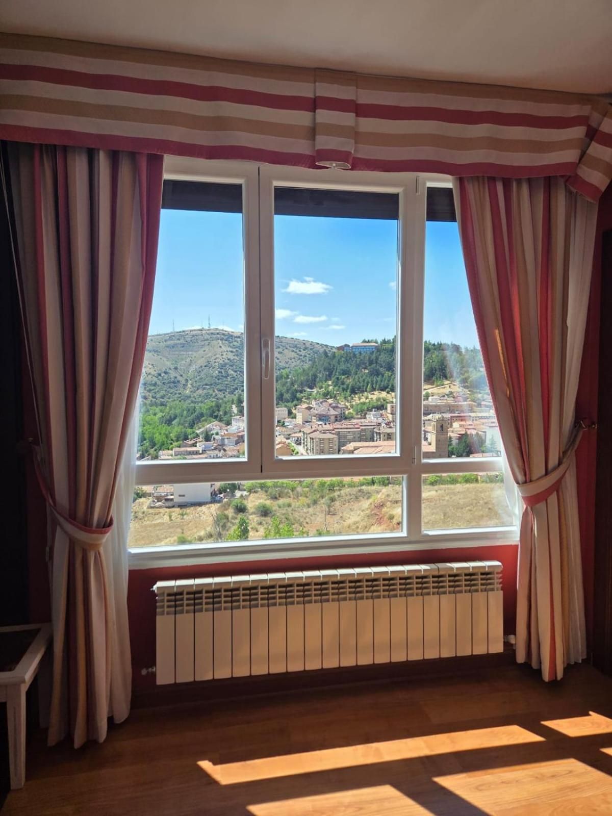 Una habitación con un ventanal grande, cortinas, un radiador y vistas a un paisaje montañoso.