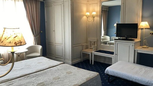 A hotel room with two beds, a television, a desk with a lamp, a chair, and a wardrobe. - Hotel Leonor Miron (Soria)