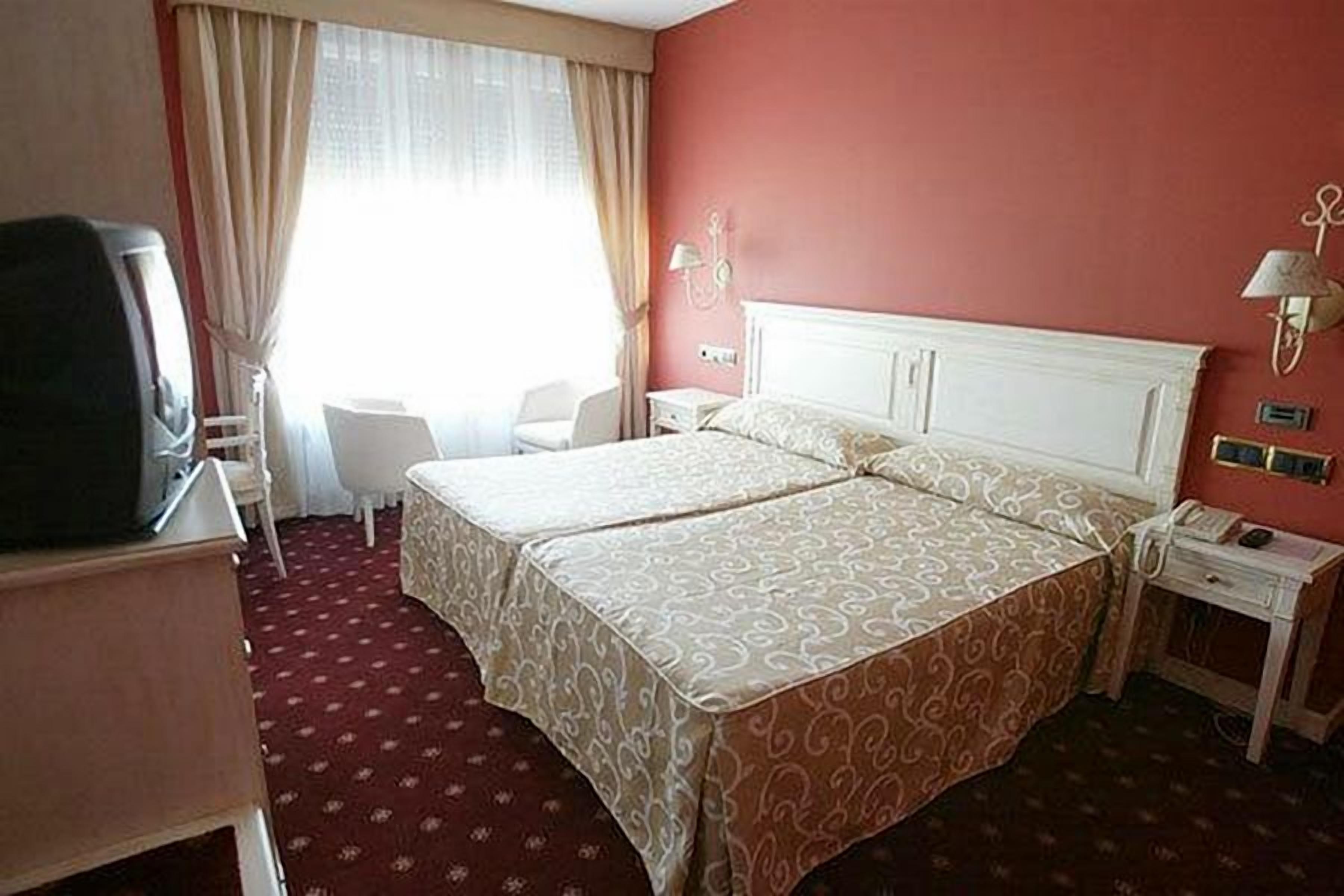 A hotel room with a double bed, a desk with a chair, a television, and a bedside table with a lamp.