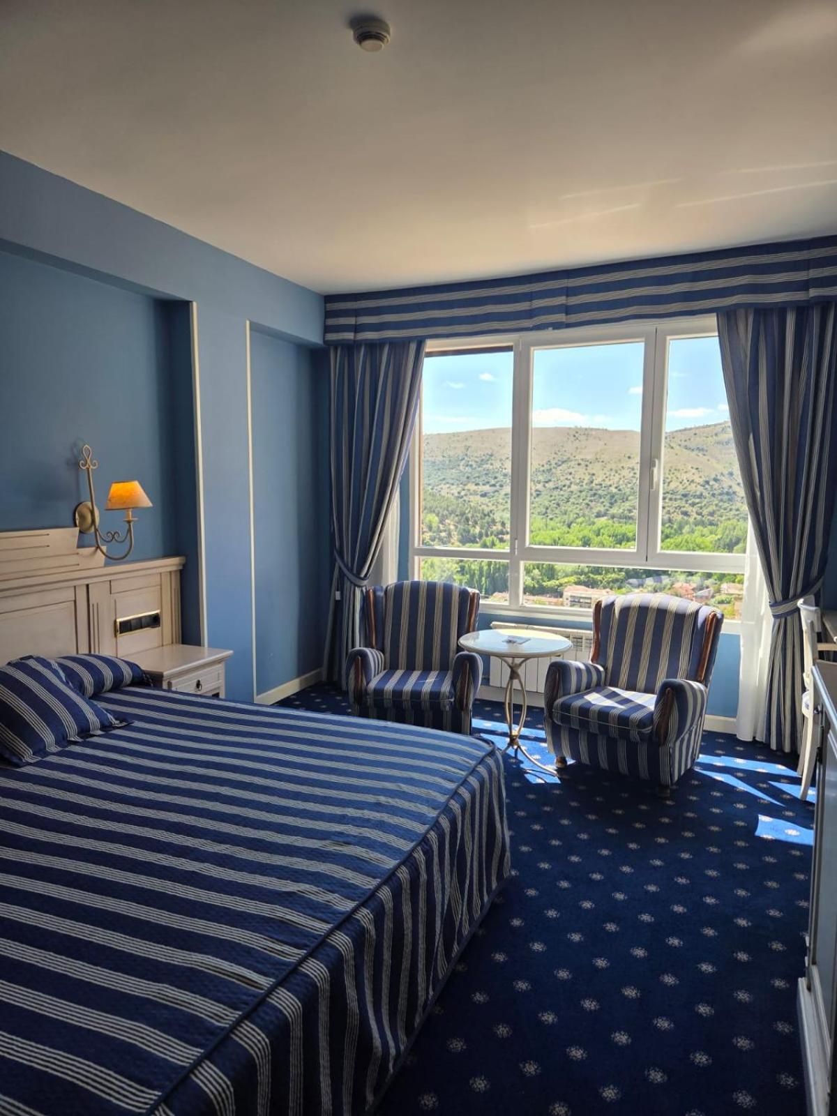 Habitación de hotel con una cama grande, dos sillones, una mesita y vistas a un paisaje montañoso.