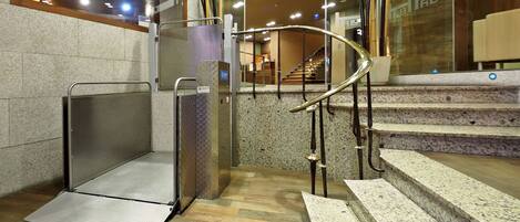 Un ascensor moderno con estructura metálica y panel de vidrio, que da acceso a una escalera con escalones de granito.