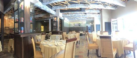 A restaurant with round tables, wicker chairs, and a high ceiling with exposed beams.