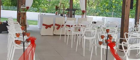 Un espacio para eventos al aire libre con sillas y mesas blancas, lazos rojos y un toldo blanco.