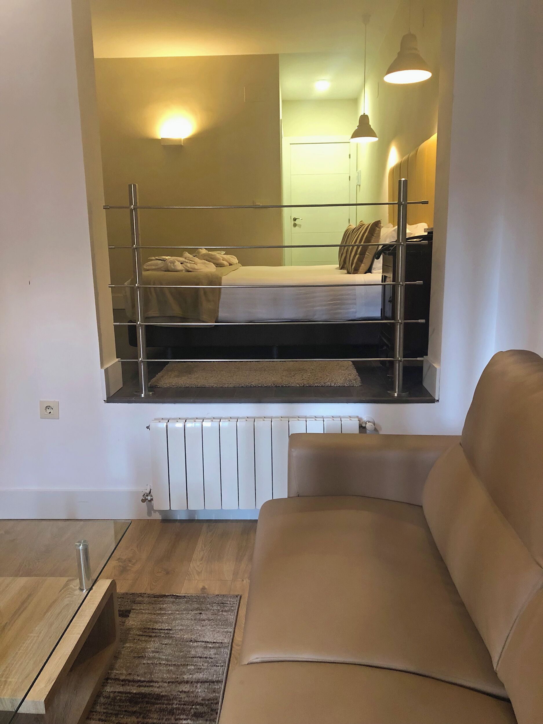 A modern living room with a sofa, a glass coffee table, and a view of a bedroom with a bed and a radiator.