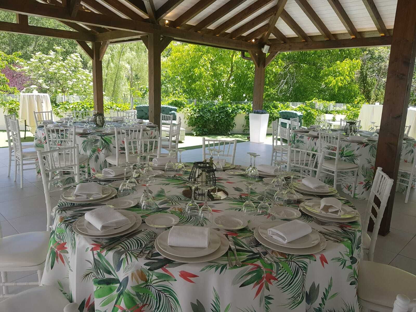 Un espacio de comedor al aire libre con mesas redondas dispuestas para una comida, rodeado de sillas blancas, bajo un pérgola de madera.
