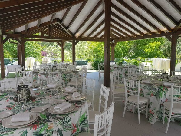 Un área de comedor al aire libre cubierta con sillas y mesas blancas, rodeada de abundante follaje.