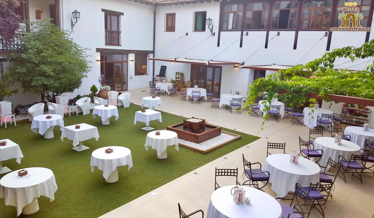 A courtyard with round tables and chairs, surrounded by a white building with multiple windows and a pergola covered with greenery.