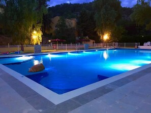 Piscina rectangular con borde de azulejos, rodeada por una valla y alumbrada por luces colgantes.