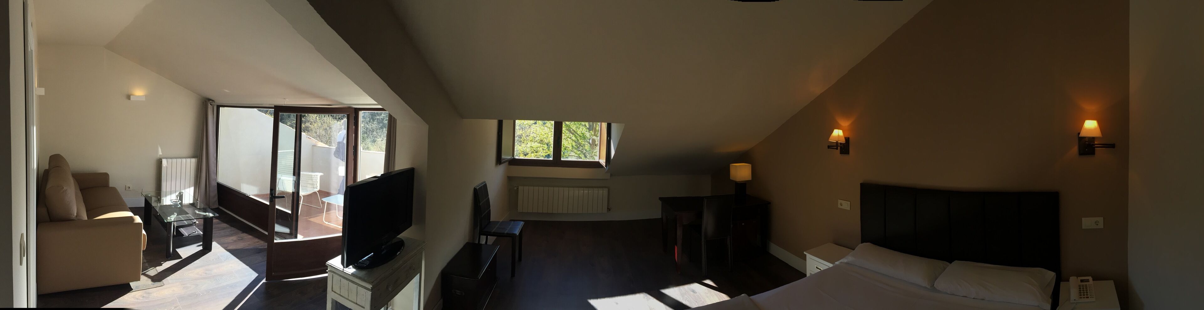 A hotel room with a bed, a TV, a desk, and a chair. There is a window providing natural light.