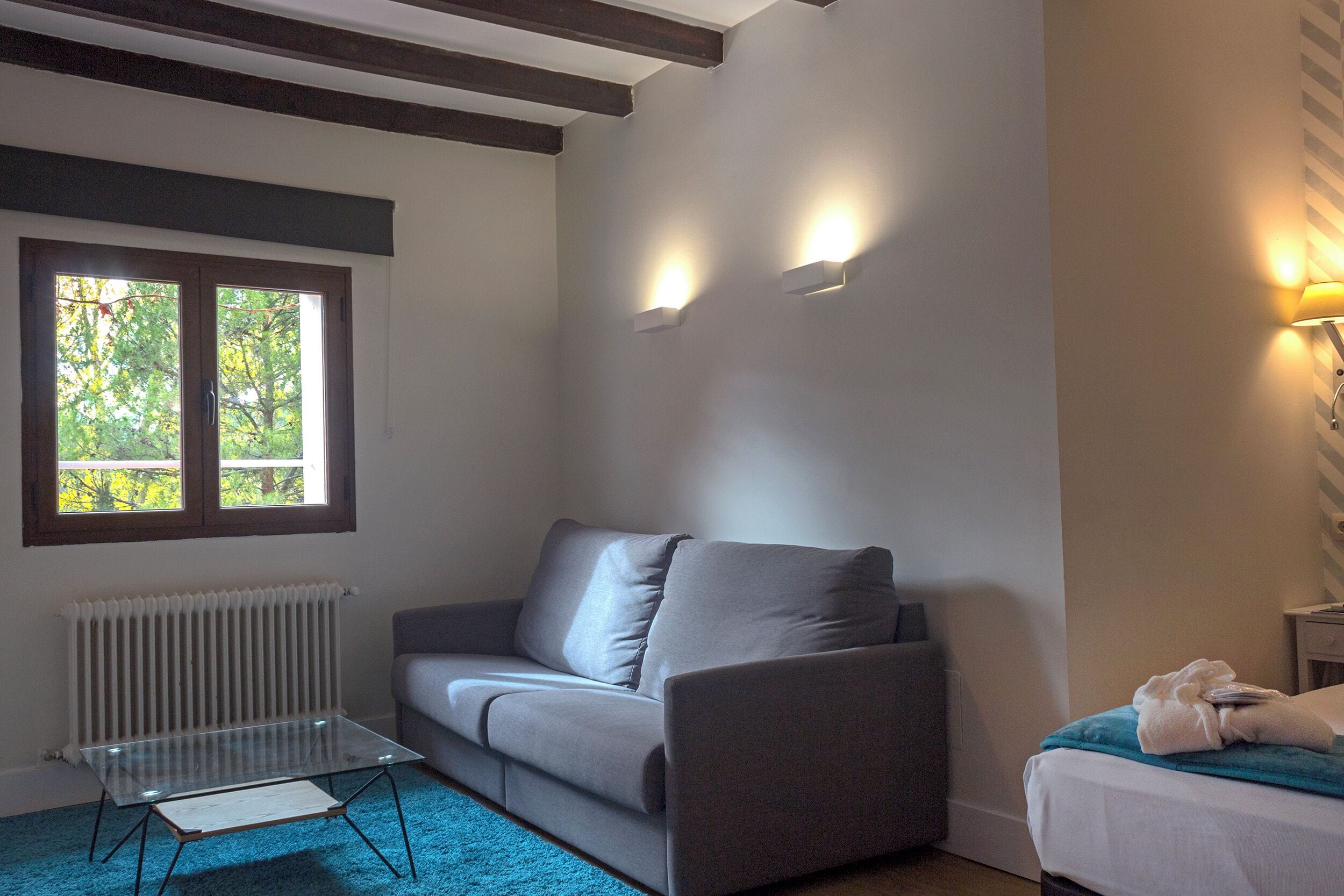Um quarto com um sofá cinzento, uma mesa de centro de vidro, um radiador, uma janela com vista para árvores e uma luminária fixada na parede.