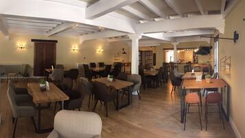 Een ruime eethoek met houten tafels en stoelen, witte balken in het plafond en wandlampen.