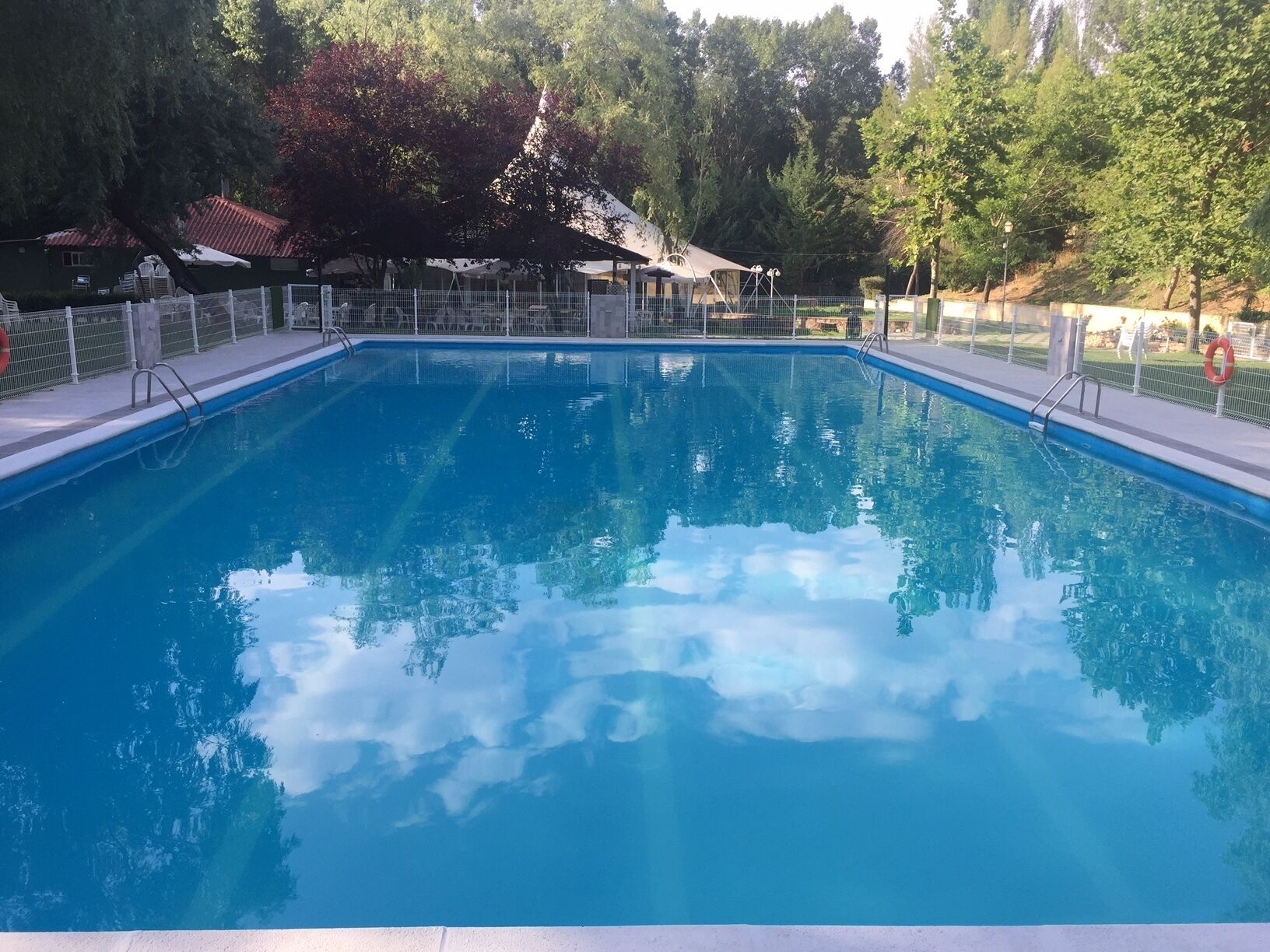 Una piscina rectangular con agua azul cristalina, rodeada por una cerca blanca y árboles.