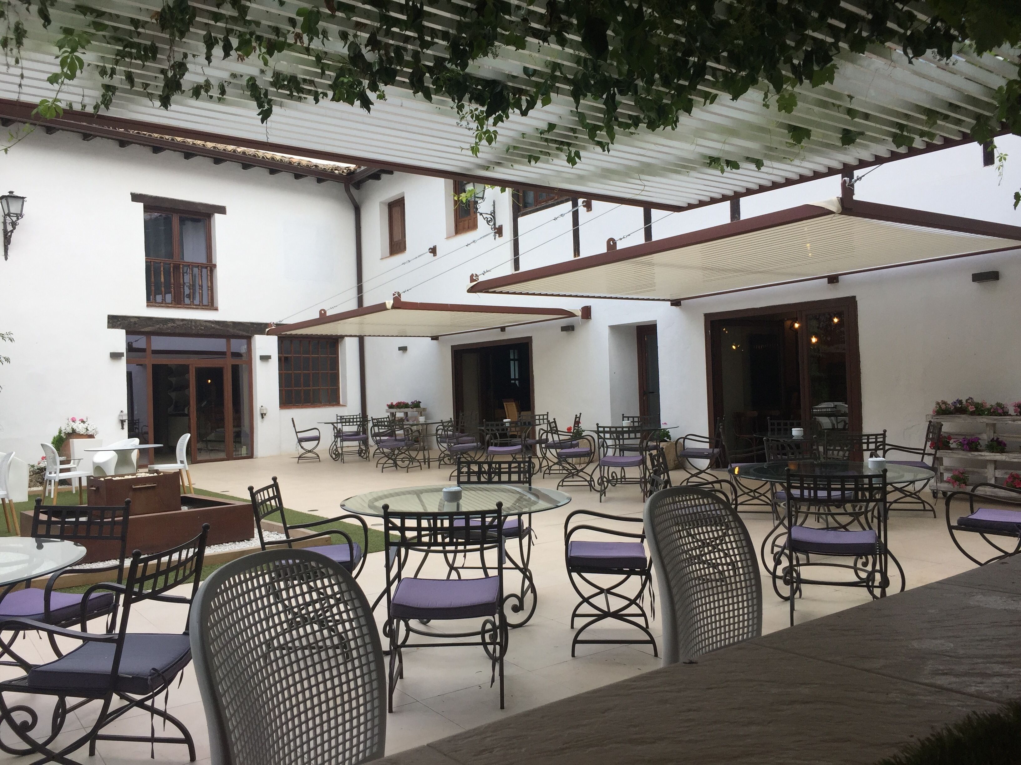 A covered outdoor seating area with tables and chairs, a white building with windows and doors, and a pergola with vines.