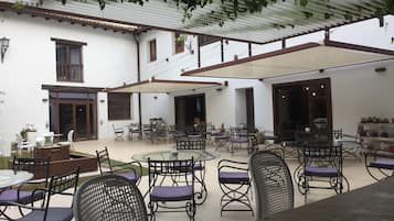 A covered outdoor seating area with tables and chairs, a white building with windows and doors, and a pergola with vines.