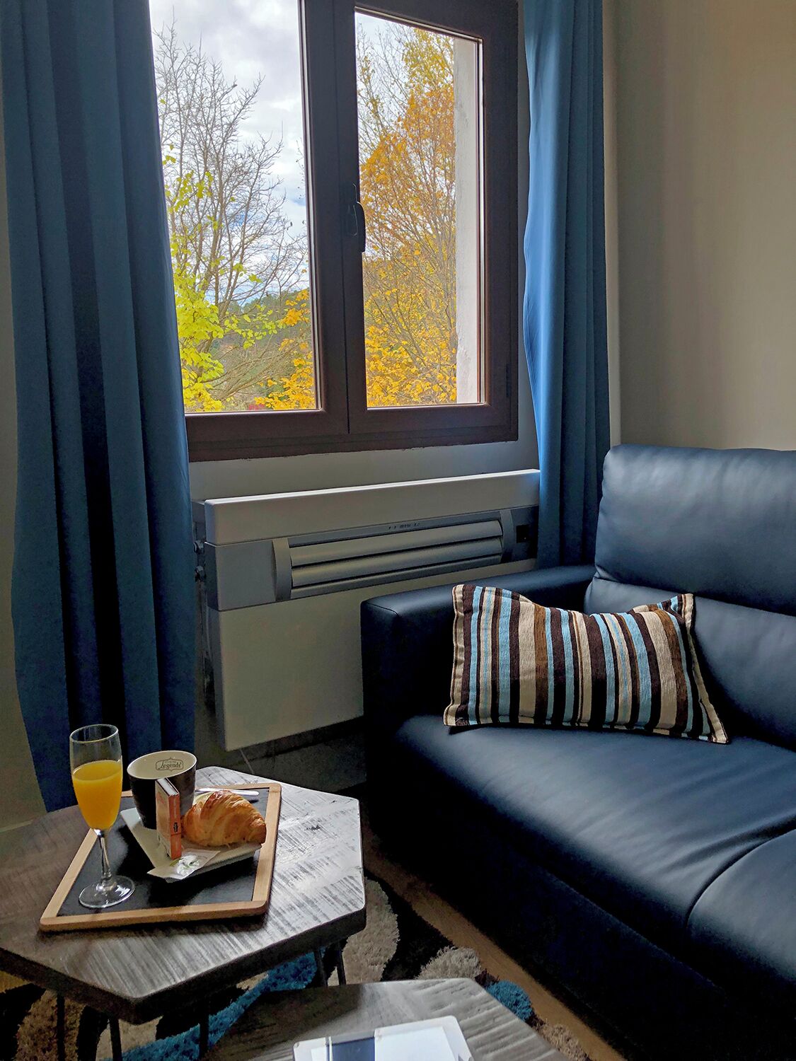 Una sala de estar acogedora con un sofá azul oscuro, una ventana con cortinas, un radiador y una mesa con una bandeja de desayuno que incluye un croissant, un vaso de zumo de naranja y una taza de café.